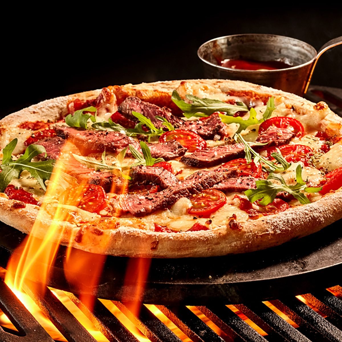 火焰伸向烤肉架上的肉类和蔬菜芝士披萨。辣酱放在铁盘上的碗里